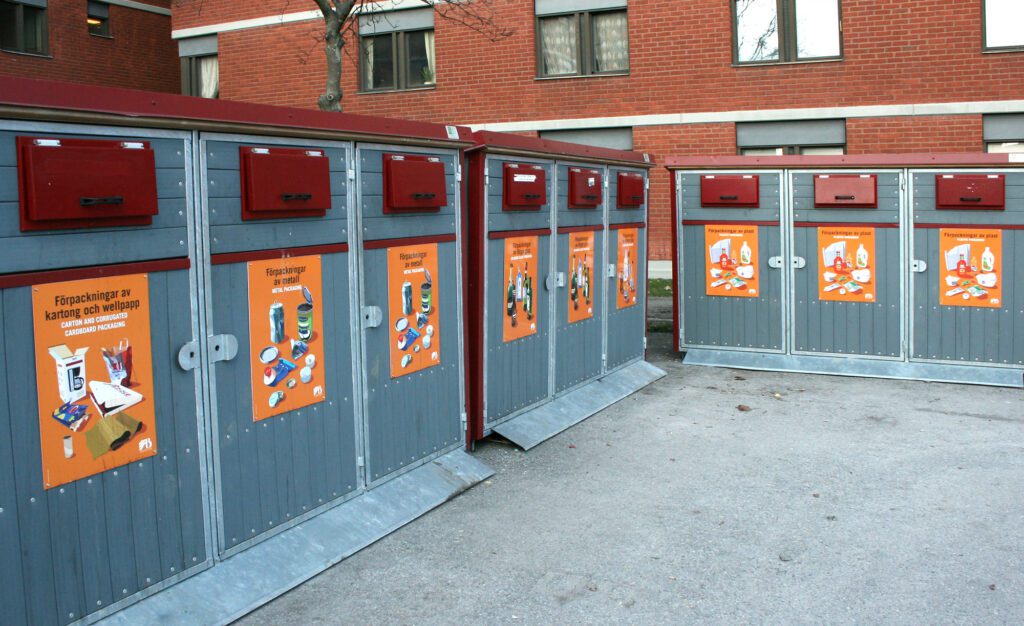 Outdoor recycling stations with grey waste cabinets and orange illustrated signs for cardboard, metal, glass and plastic packaging.